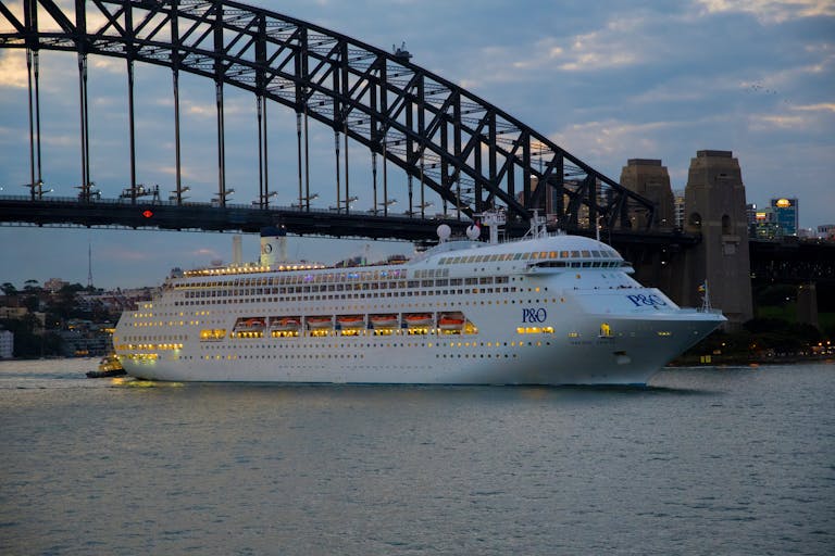 Elegant cruise ship navigates under Sydney's iconic Harbour Bridge at twilight.